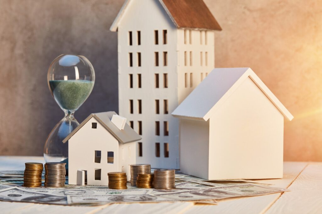 houses models, coins and cash, hourglass on white wooden table with sunlight, real estate concept